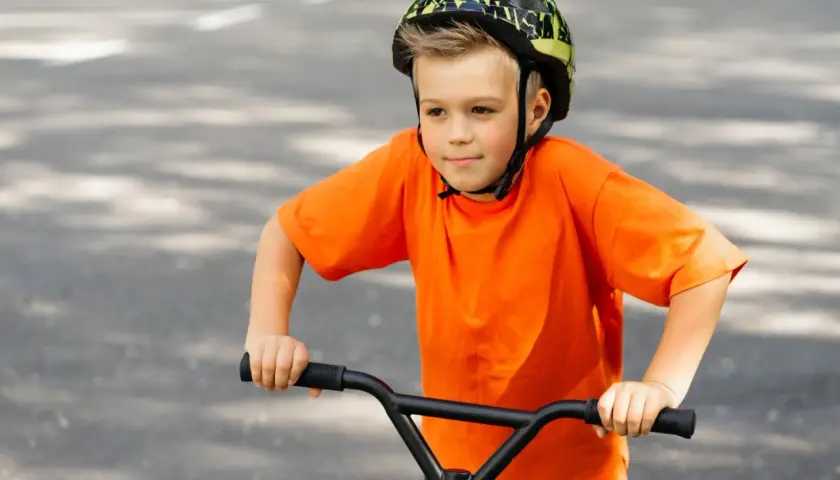 Chłopiec w kasku na rowerze, ubrany na pomarańczowo, z uśmiechem na twarzy. Czy kask na rower jest obowiązkowy? Zawsze warto go mieć!