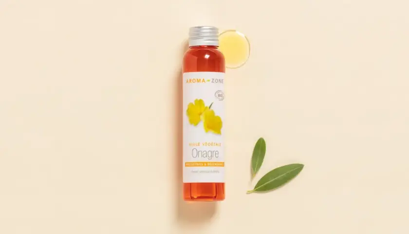 Flacon d'huile d'onagre Aroma-Zone, aux multiples bienfaits pour la peau et les cheveux. Une goutte d'huile dorée et deux feuilles vertes complètent la scène.