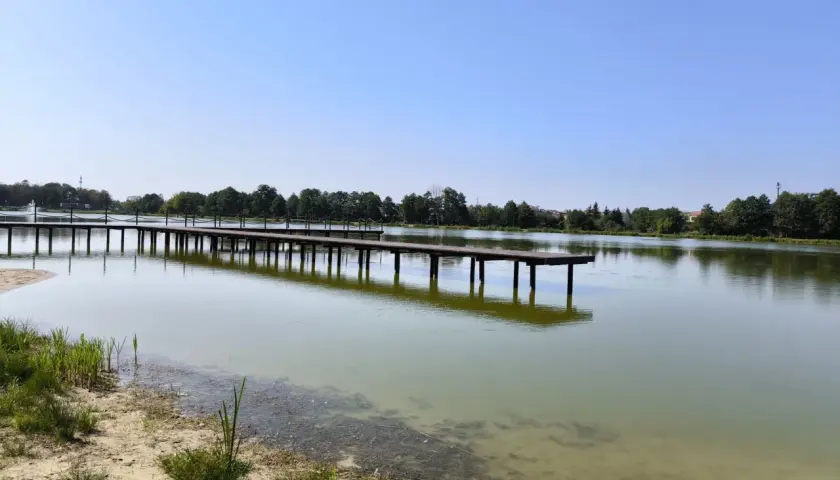 Drewniane molo w Krasnosielcu w letni, słoneczny dzień. Woda spokojna, odbija niebo i drzewa na horyzoncie.