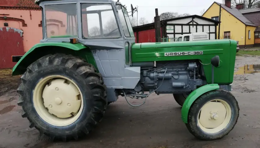 Zielony Ursus c355 z szarą kabiną, gotowy do pracy na błotnistym podwórku.