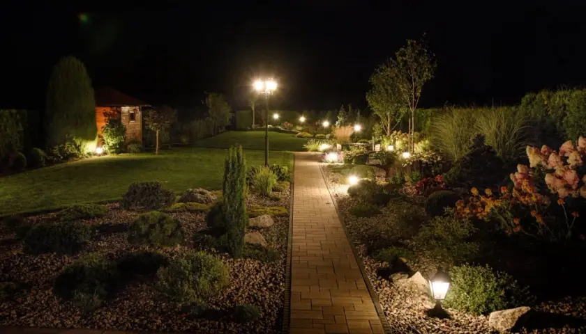 Piękna ścieżka z kostki brukowej prowadzi przez ogród, który nocą rozświetla nastrojowe oświetlenie. Inspiracje dla Twojego ogrodu.