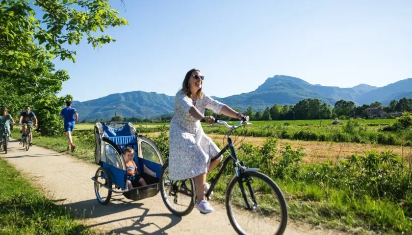 Disfrutando de un día soleado en la ruta el carrilet. Una mujer pedalea con un niño en un remolque, mientras otros ciclistas y un corredor disfrutan del paisaje montañoso.