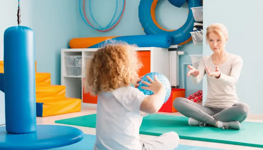 Dziecko z kręconymi włosami bawi się piłką, a kobieta instruuje je w sali do terapii SI.
