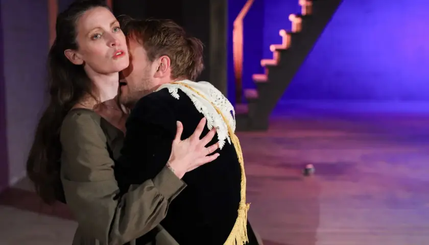 En kvinna och en man omfamnar varandra passionerat på en scen. Bakom dem syns en trappa och lila belysning. Slutet gott allting gott.