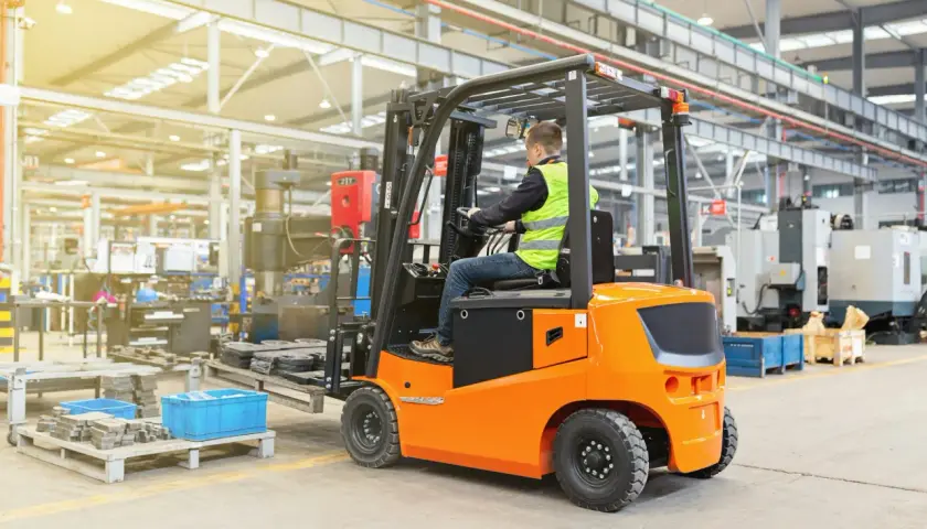 Ein Mann in Warnweste fährt einen orangefarbenen Gabelstapler in einer Fabrikhalle. Die Nutzungsdauer des Gabelstaplers ist entscheidend für die Effizienz.