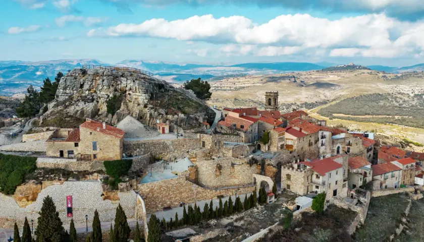 Vista aérea de un pueblo de casas de piedra con tejados rojos, anidado en la ladera de la roca del corb, bajo un cielo azul con nubes.