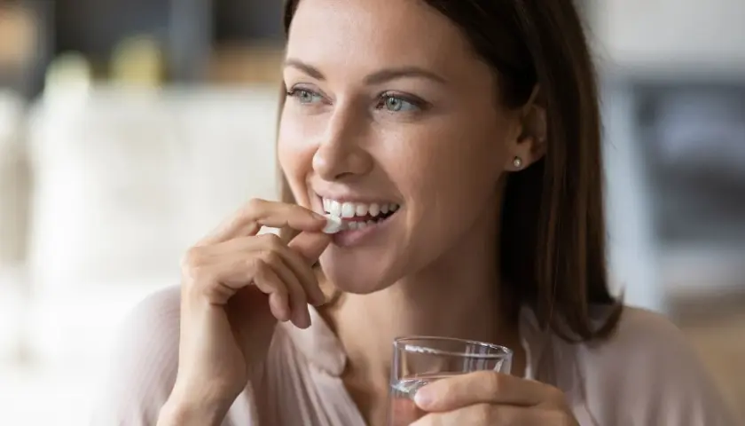Femme souriante prenant un comprimé blanc, rappelant l'importance de ne pas associer le metronidazole alcool. Verre d'eau à la main.