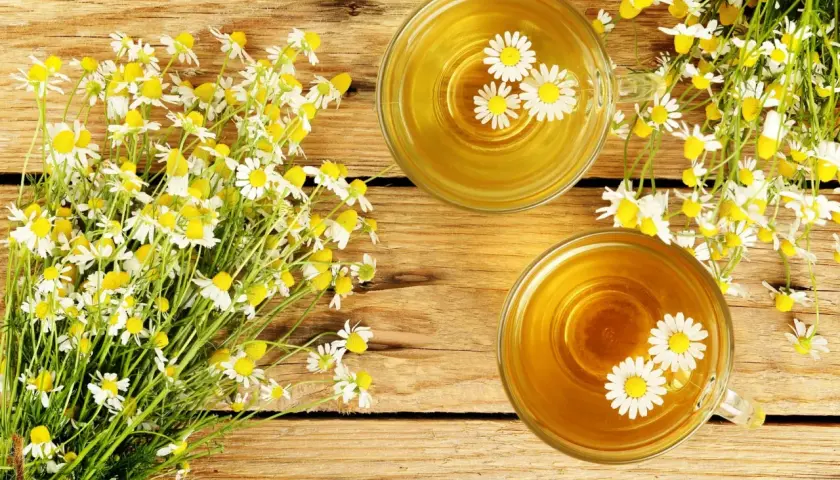 Due tazze di tisana dorata con fiori di camomilla galleggianti, accanto a un mazzo di fiori freschi. Rimedio naturale per il bruciore di stomaco.