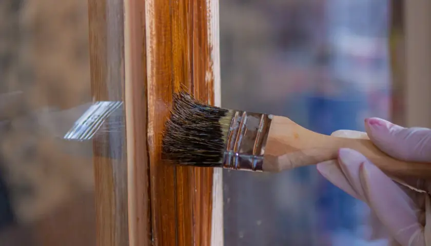 Eine Hand mit Handschuh streicht mit einem Pinsel einen braunen Holzrahmen. Das Holzfenster erstrahlt bald in neuem Glanz.