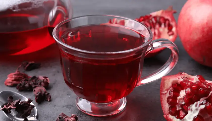 Aromatyczna herbata granat w szklance, obok świeże owoce granatu i suszone kwiaty hibiskusa.