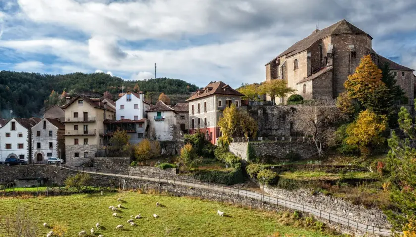 Casas de piedra y tejados rojos se asoman en el **valle de Ansó**, con ovejas pastando en un prado verde y un cielo nublado.