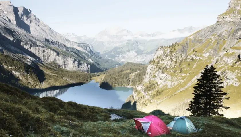 Two tents are set up on a grassy slope overlooking a serene lake in the Swiss Alps. This is a perfect spot for swiss alps hiking and camping.