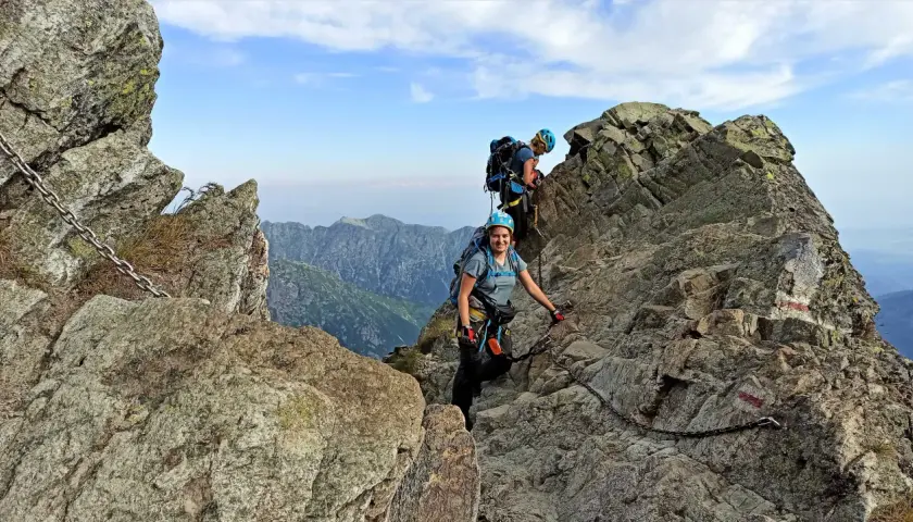 Dwoje turystów wspina się po skalistych tatry wysokie szlaki, korzystając z łańcuchów. Widok na góry i błękitne niebo.