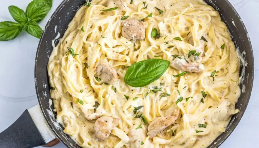 Un cremoso plato de pasta alfredo con trozos de pollo y hojas de albahaca fresca en una sartén.