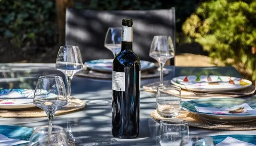 Une bouteille de vin rouge est prête pour un apéro dinatoire en extérieur, entourée de verres et d'assiettes sur une table de jardin.
