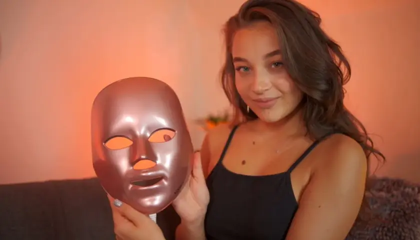 En ung kvinna håller upp en roséguld LED-mask för ansiktet, redo för en led-ljus behandling ansikte.