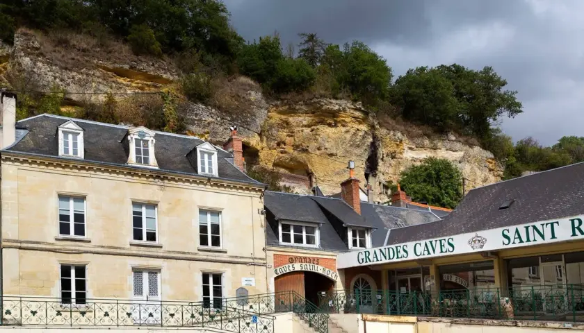 La maison Lacheteau, avec ses façades en pierre et ses toits d'ardoise, se dresse au pied d'une falaise rocheuse.