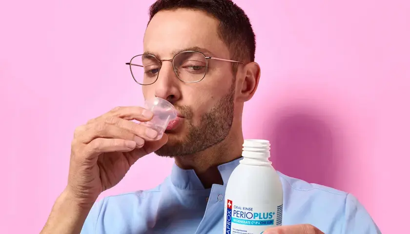 Uomo con occhiali beve collutorio da un bicchierino. Il flacone di Curasept gel va risciacquato.