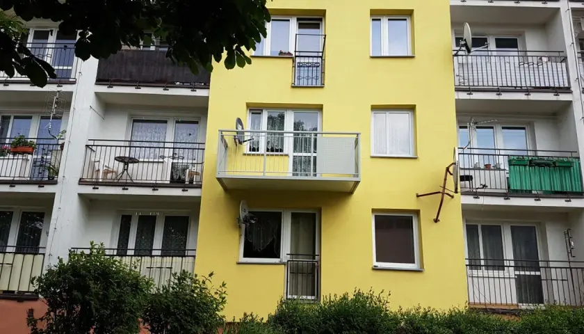 Żółty blok z dobudową balkonu na słupach. Widać okna, balustrady i anteny satelitarne.
