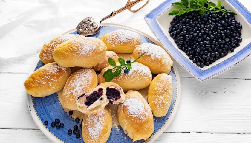 Pyszne jagodzianki posypane cukrem pudrem, obok miseczka świeżych jagód. Idealne na lato, zgodnie z najlepszym jagodzianki przepis.