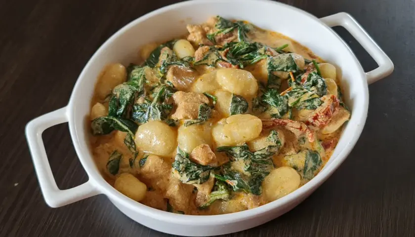 Pyszne gnocchi z kurczakiem, szpinakiem i suszonymi pomidorami w kremowym sosie. Danie podane w białej owalnej misce.