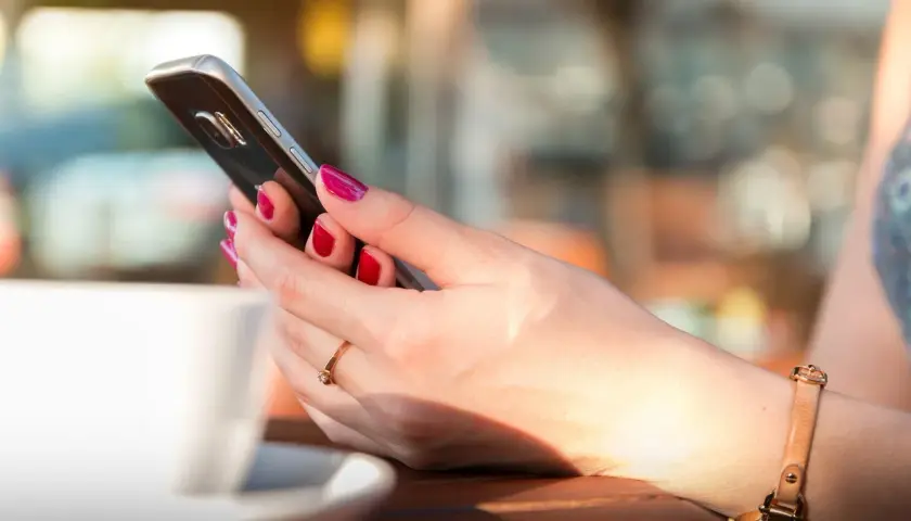 Dłoń z pomalowanymi paznokciami trzyma smartfon, obok biała filiżanka. Długą żywotność baterii w telefonie doceni każda użytkowniczka.