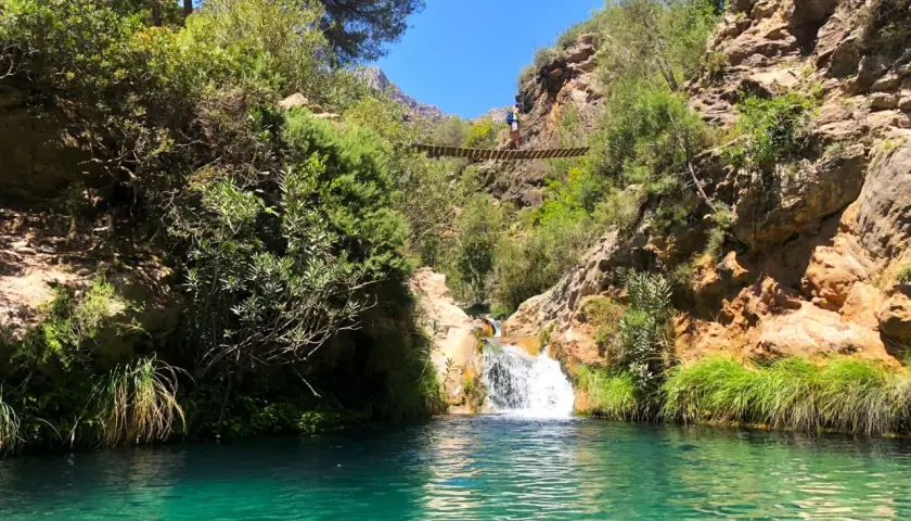 Un puente colgante cruza un **río verde** en Granada, con una cascada y vegetación exuberante.