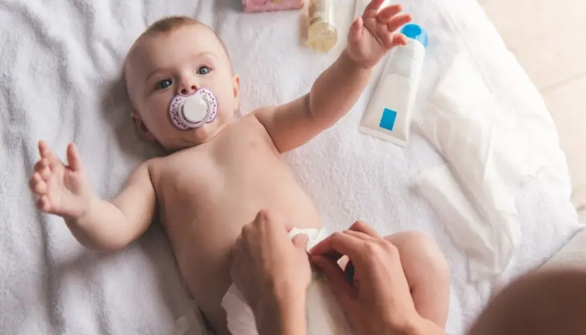 Chłopiec z smoczkiem leży na ręczniku, a ręce mamy dokonują **przewijania chłopca**. Obok kosmetyki i chusteczki.