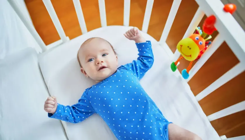 Urocze, 10 tygodniowe dziecko w niebieskim pajacyku leży w łóżeczku obok kolorowej zabawki.