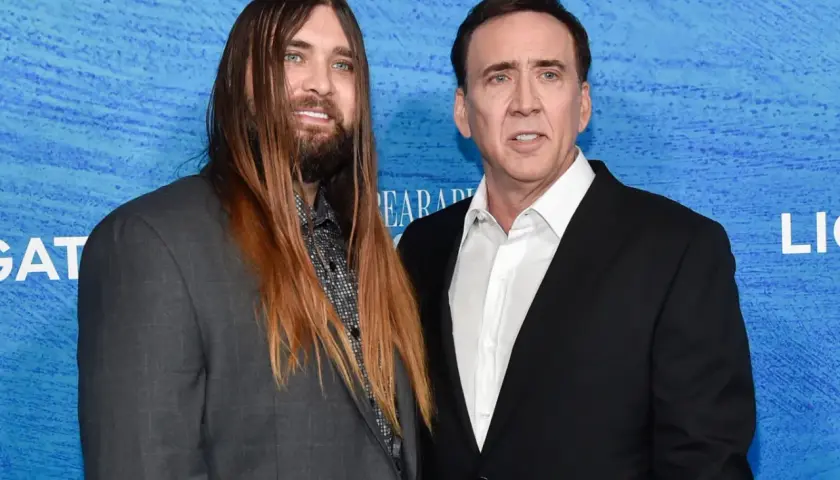 Weston Coppola Cage z długimi włosami i Nicolas Cage w czarnym garniturze pozują do zdjęcia na niebieskim tle.