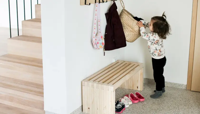 Dziecko wiesza torbę na haczyku nad drewnianą ławką. Prosty projekt, jak zrobic ławkę, idealny do przedpokoju.