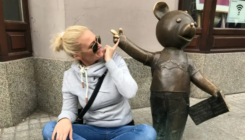 Kobieta w okularach przeciwsłonecznych i szarej bluzie bawi się z pomnikiem misia, który trzyma tabliczkę. To część szlaku łodzi bajkowej.