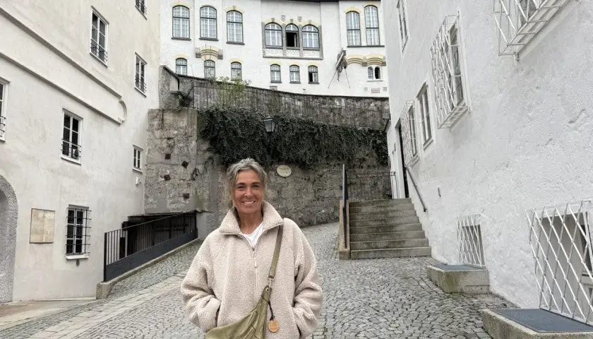 Eine Frau steht lächelnd auf einem Kopfsteinpflasterweg zwischen alten weißen Häusern. Sie könnte eine Pilgerin sein, die auf ihrer Reise neue Orte entdeckt.
