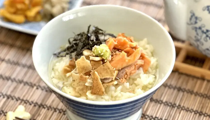 A delicious ochazuke recipe featuring rice, flaked salmon, nori, and wasabi in a blue-striped bowl, with a teapot in the background.