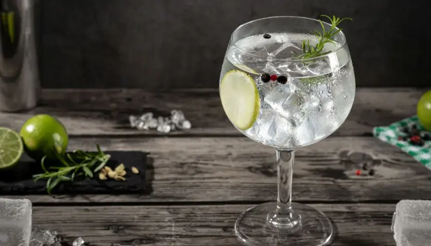Orzeźwiający gin and tonic z plasterkiem limonki, gałązką rozmarynu i ziarnami pieprzu, na drewnianym blacie.