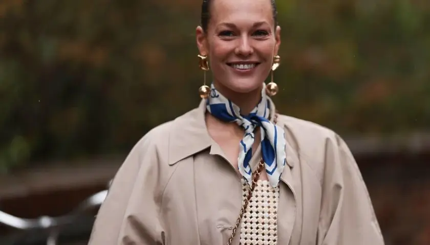 Uśmiechnięta kobieta w beżowym płaszczu i stylowej apaszce z niebieskimi wzorami. Inspiracja, jak nosić apaszkę, by dodać elegancji.