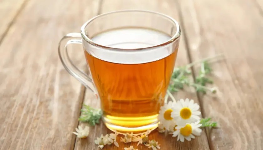 Ein Glas mit goldenem Kamillentee steht auf Holz. Kleine Kamillenblüten schmücken die Szene. Perfekt, um kamillentee kalt trinken.