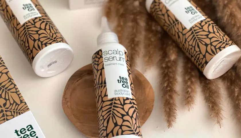 Flera flaskor med "tea tree oil" hårvårdsprodukter, inklusive en "scalp serum" treatment mask, arrangerade med torkade grässtrån.