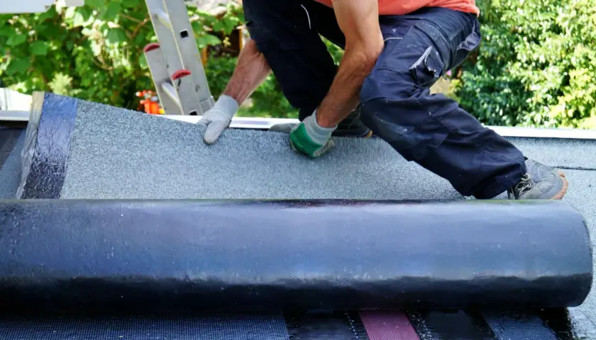 Handwerker klebt Dachpappe. Mit Handschuhen wird die Bitumenbahn sorgfältig ausgerollt und befestigt, um das Dach wetterfest zu machen.