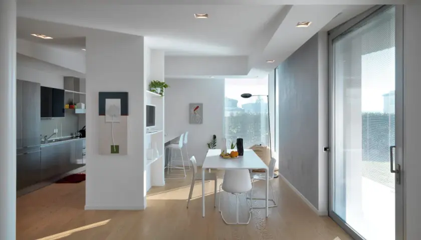 Interno moderno con cucina a vista, zona pranzo e un'ampia vetrata che sembra allargare una finestra in condominio, inondando di luce naturale.