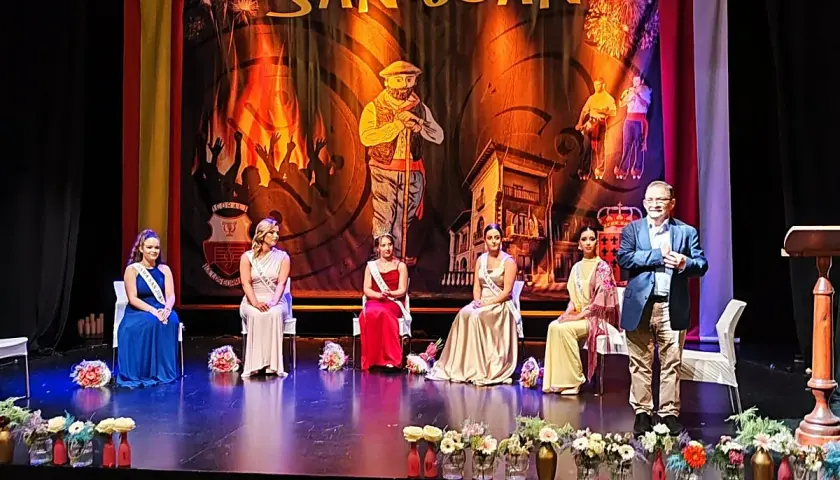 En el teatro Los Corrales de Buelna, cinco damas de honor y un presentador esperan en el escenario bajo un telón de San Juan.