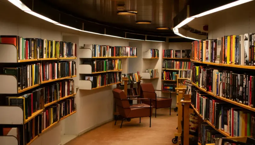 Wnętrze biblioteki z regałami pełnymi książek. Wygodne fotele zapraszają do lektury, a każdy tom czeka na swojego bibliotekarza.