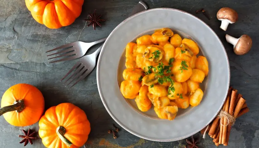 Pyszne gnocchi z kremowym sosem dyniowym, ozdobione natką pietruszki. Obok talerza małe dynie i przyprawy.