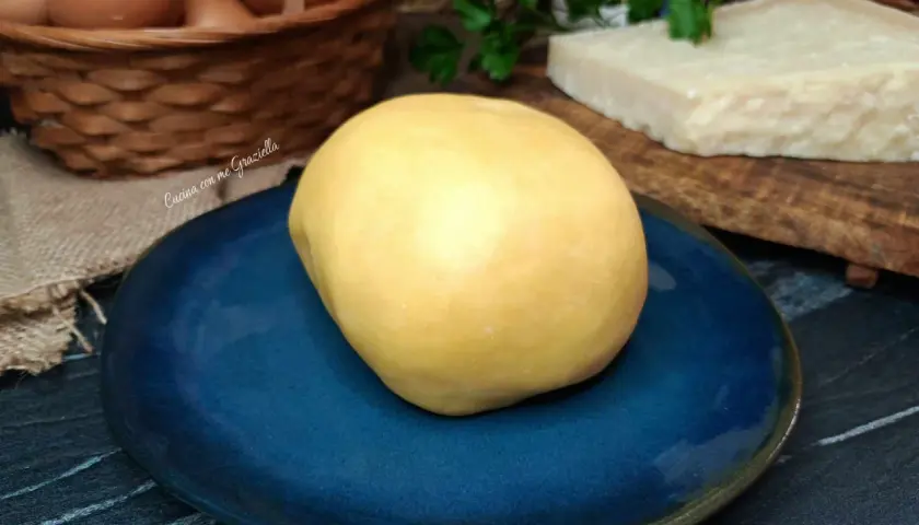 Impasto giallo per frolla salata su piatto blu, con uova e parmigiano sullo sfondo.