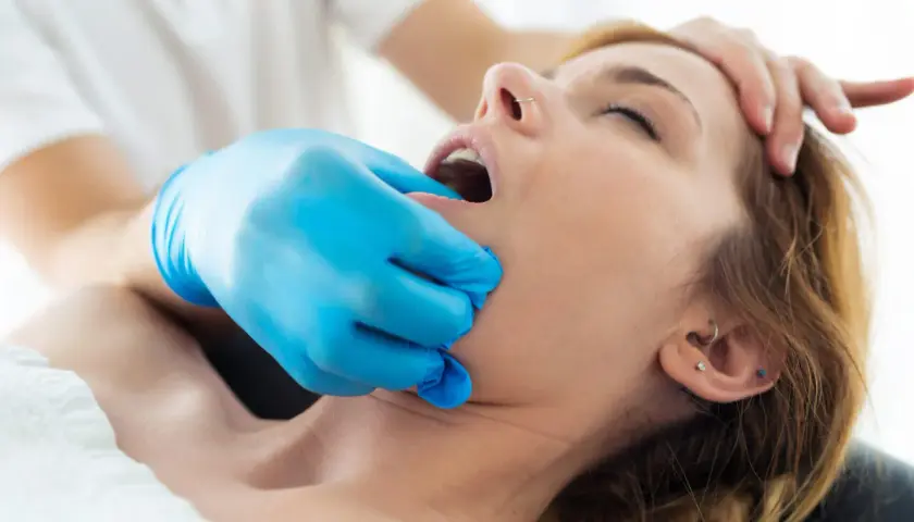 Fizjoterapeuta w niebieskiej rękawiczce delikatnie bada szczękę pacjentki, która cierpi na **szczekoscisk**.