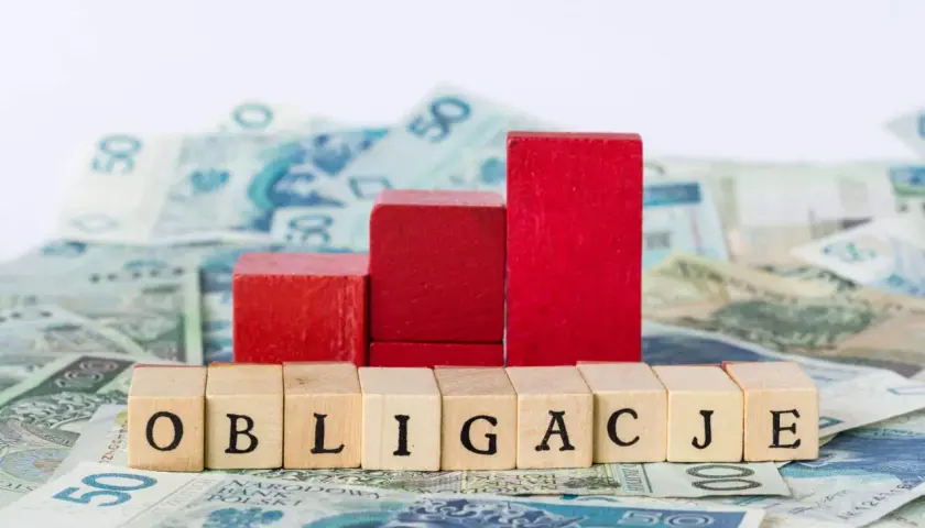 Drewniane klocki z napisem "OBLIGACJE" na tle polskich banknotów. Czerwone klocki tworzą wykres wzrostu.