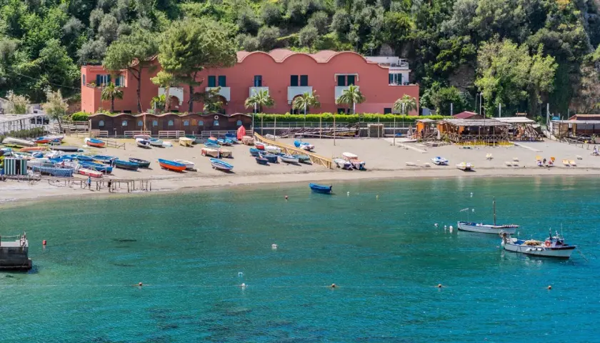 Plage de Vico Equense avec des bateaux colorés sur le sable et dans l'eau turquoise. Bâtiment rose en arrière-plan.
