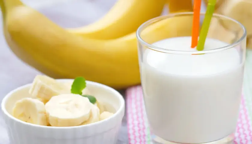 Szklanka pysznego mleka bananowego z dwoma słomkami, obok miseczka z pokrojonymi bananami i całe banany w tle.