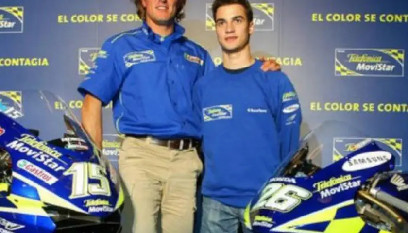 Sete Gibernau, con su edad, posa junto a un joven piloto, ambos con ropa de equipo azul, frente a motocicletas de competición.