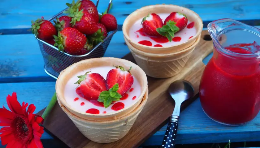 Panna cotta w wafelkach z truskawkami i miętą, obok dzbanuszek z musem truskawkowym.
